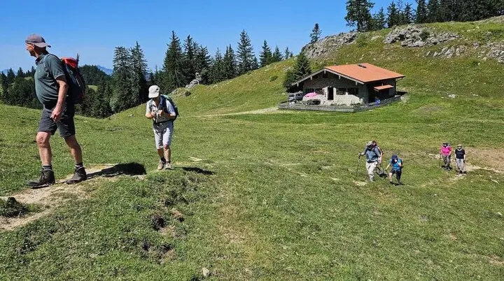Fünf ältere Wanderer steigen bei sonnigem Wetter einen grasbewachsenen Hügel hinauf, Richtung Berghütte mit rotem Dach. | © DAV Markt Schwaben | Foto Hubert Inhofer