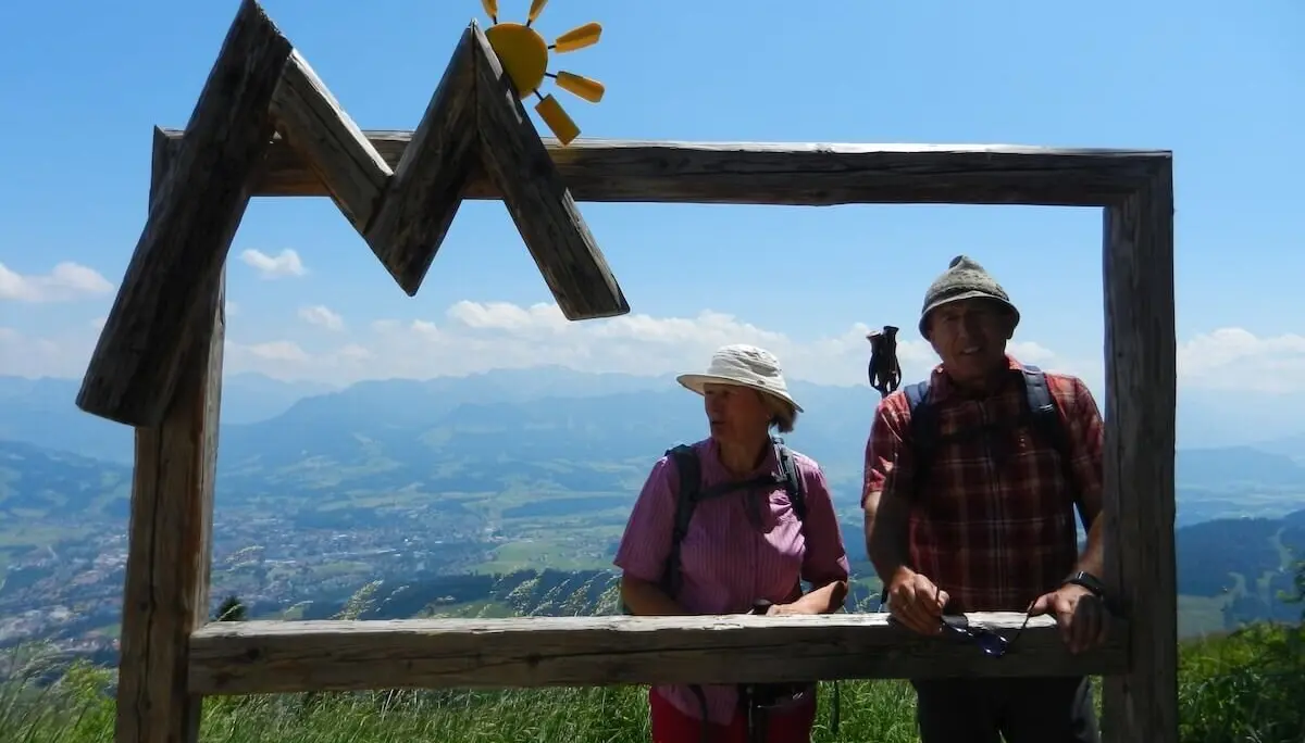 Zwei Wandernde posieren hinter einem Holzrahmen in Bergform mit Sonne, im Hintergrund das Allgäu-Panorama bei klarem Himmel. | © DAV Markt Schwaben