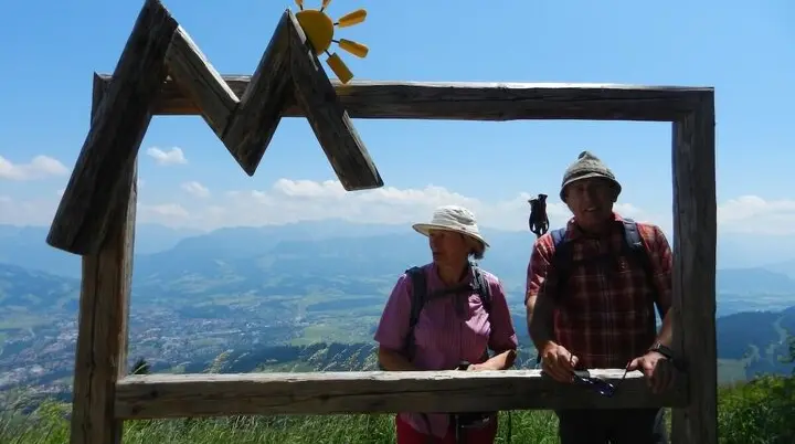 Zwei Wandernde posieren hinter einem Holzrahmen in Bergform mit Sonne, im Hintergrund das Allgäu-Panorama bei klarem Himmel. | © DAV Markt Schwaben