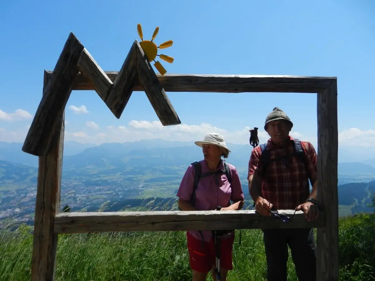 Zwei Wandernde posieren hinter einem Holzrahmen in Bergform mit Sonne, im Hintergrund das Allgäu-Panorama bei klarem Himmel. | © DAV Markt Schwaben