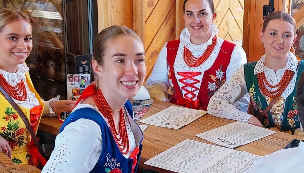 Vier Personen in bunter polnischer Festtracht sitzen an einem Holztisch in rustikalem Raum und blättern durch Speisekarten. | © DAV Markt Schwaben | Foto Lutz Gründel
