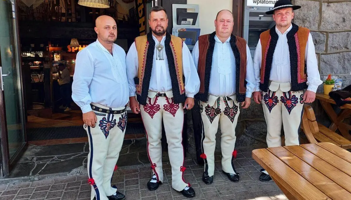 Vier Männer in bestickter Goral-Tracht stehen vor einem rustikalen Gebäude mit Bankomat-Schild und Holzbänken im Hintergrund. | © DAV Markt Schwaben | Foto Lutz Gründel