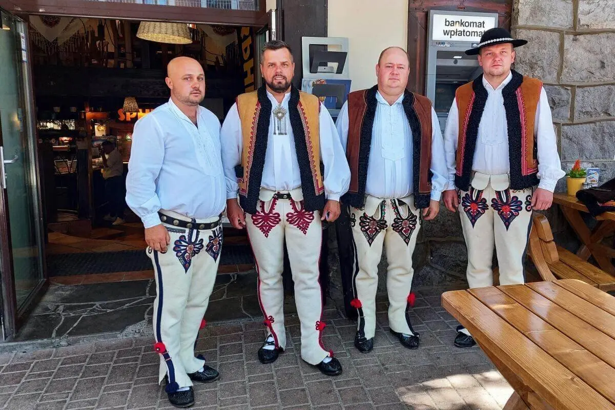 Vier Männer in bestickter Goral-Tracht stehen vor einem rustikalen Gebäude mit Bankomat-Schild und Holzbänken im Hintergrund. | © DAV Markt Schwaben | Foto Lutz Gründel