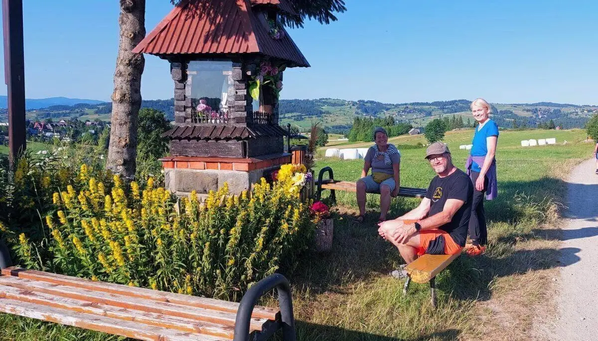 Drei Personen ruhen an einem blumengeschmückten Wegschrein mit rotem Dach; im Hintergrund Felder, Hügel und blauer Himmel. | © DAV Markt Schwaben | Foto Lutz Gründel