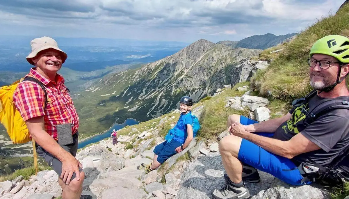 Drei Wandernde sitzen auf Felsen oberhalb eines Bergsees; im Hintergrund steile Hänge, Wolken und weite Aussicht ins Tal. | © DAV Markt Schwaben | Foto Sabine Hainz