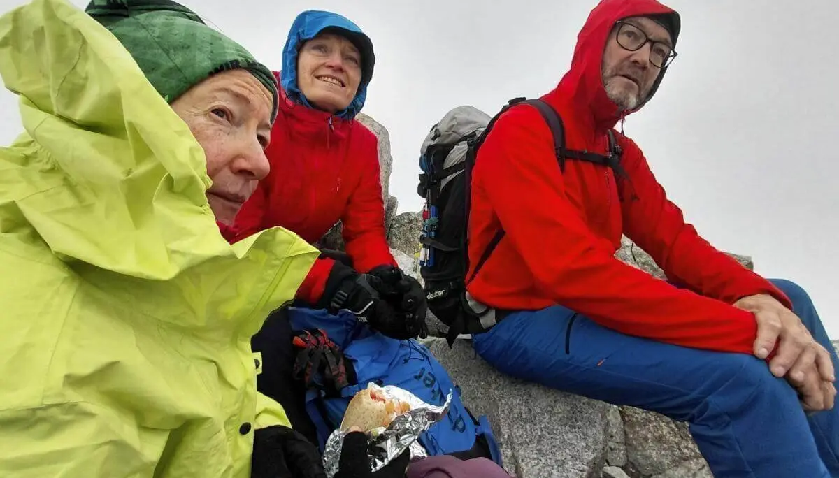 Drei Wandernde sitzen auf Felsen in nebliger Höhe; sie tragen bunte Regenjacken und essen bei windigem Wetter eine Brotzeit. | © DAV Markt Schwaben | Foto Sabine Hainz