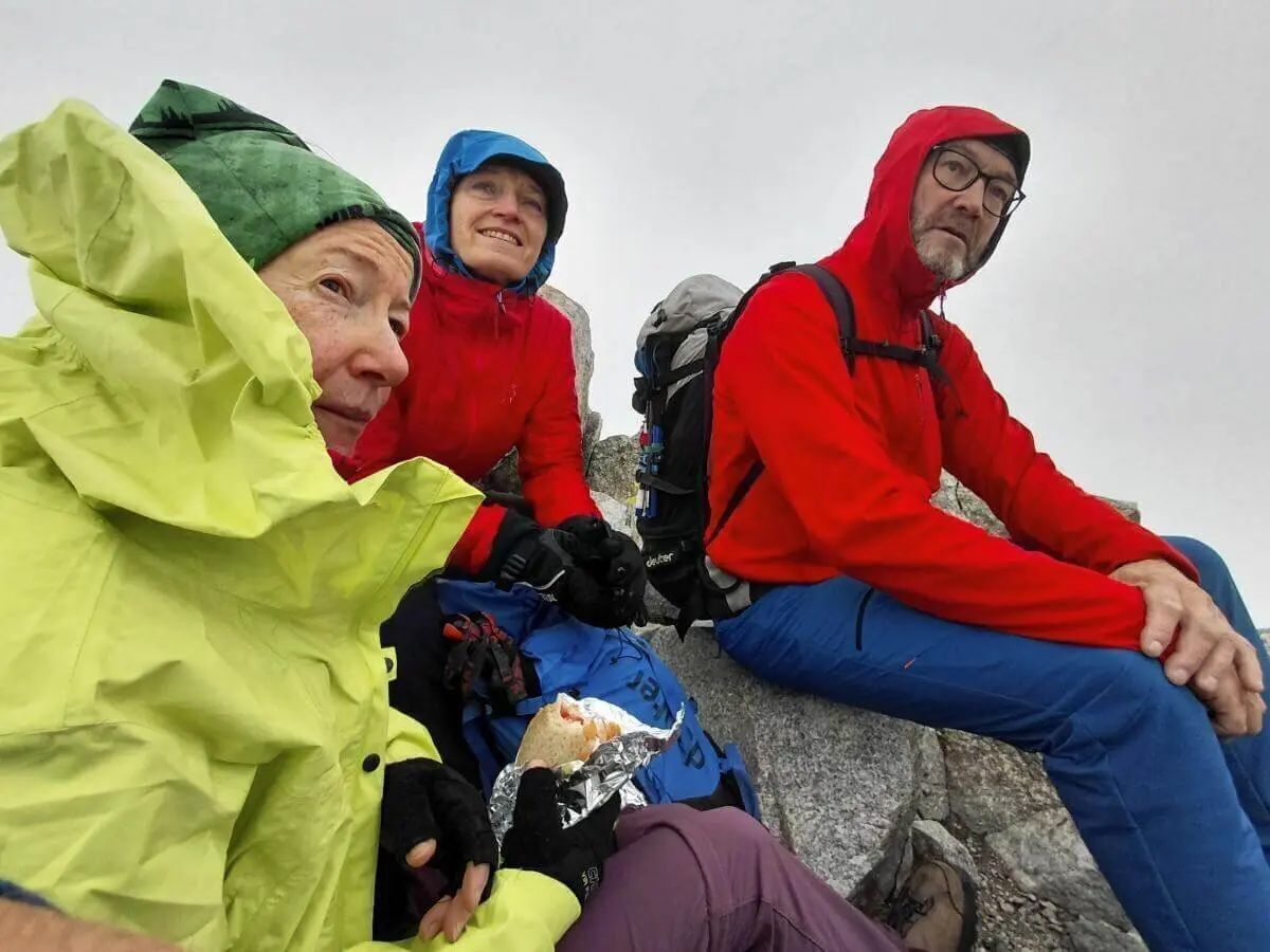 Drei Wandernde sitzen auf Felsen in nebliger Höhe; sie tragen bunte Regenjacken und essen bei windigem Wetter eine Brotzeit. | © DAV Markt Schwaben | Foto Sabine Hainz