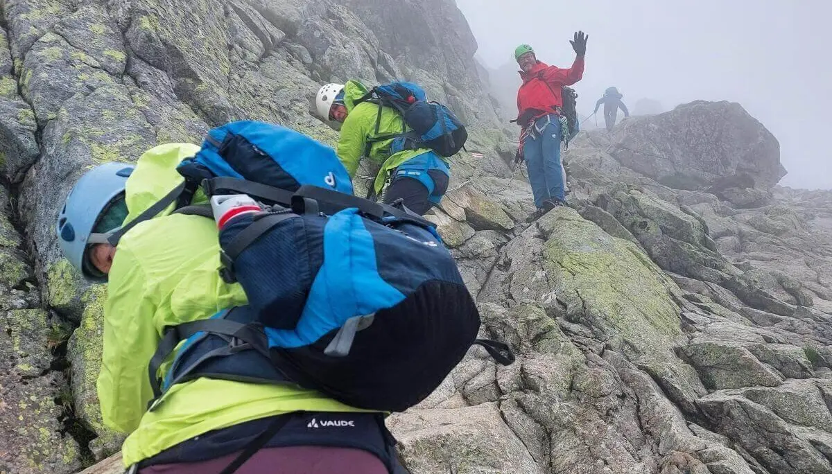 Mehrere Kletternde steigen mit Helm und Seil durch nebliges Felsgelände; eine Person winkt, andere sichern sich am steilen Hang. | © DAV Markt Schwaben | Foto Lutz Gründel