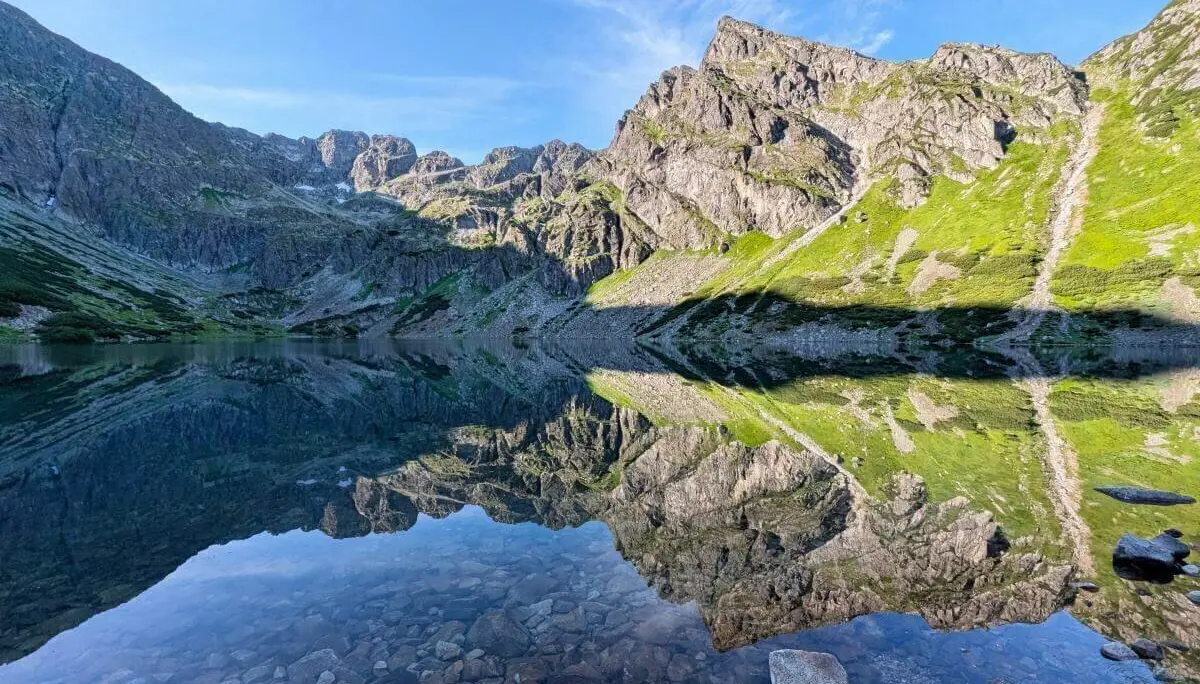 Ein klarer Bergsee spiegelt Felsen, Himmel und Wolken; im Vordergrund sind Steine sichtbar, dahinter grüne Hänge und Gipfel. | © DAV Markt Schwaben | Foto Michael Huber