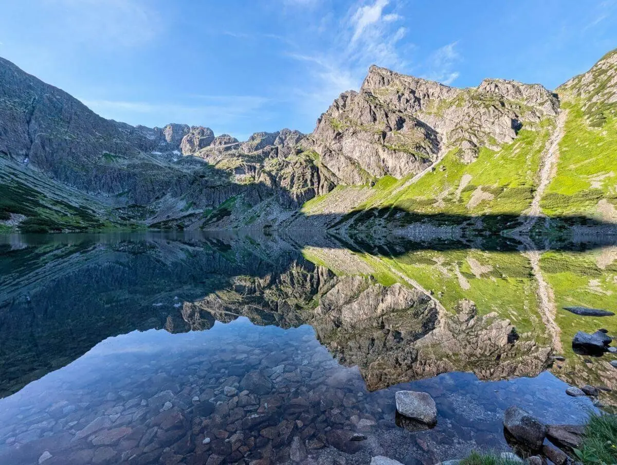 Ein klarer Bergsee spiegelt Felsen, Himmel und Wolken; im Vordergrund sind Steine sichtbar, dahinter grüne Hänge und Gipfel. | © DAV Markt Schwaben | Foto Michael Huber