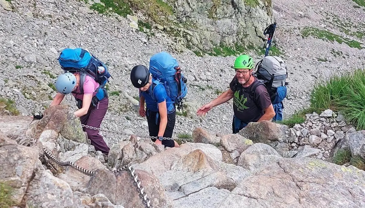 Vier Wandernde steigen mit Helm und Rucksack über steilen Felsgrat; sie sichern sich mit Ketten im nebligen Hochgebirge. | © DAV Markt Schwaben | Foto Lutz Gründel