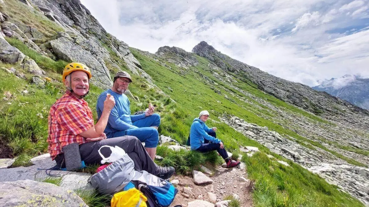 Drei Wandernde sitzen auf felsigem Hang; zwei zeigen Daumen hoch, im Hintergrund steile Felsen, Gras und wolkiger Himmel. | © DAV Markt Schwaben | Foto Michaela Haaß