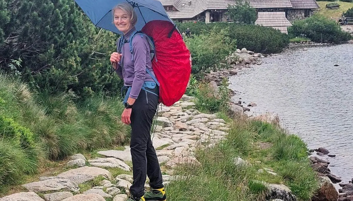 Eine Person mit rotem Rucksack und blauem Schirm wandert am See entlang; im Hintergrund ein Holzhaus. | © DAV Markt Schwaben | Foto Lutz Gründel
