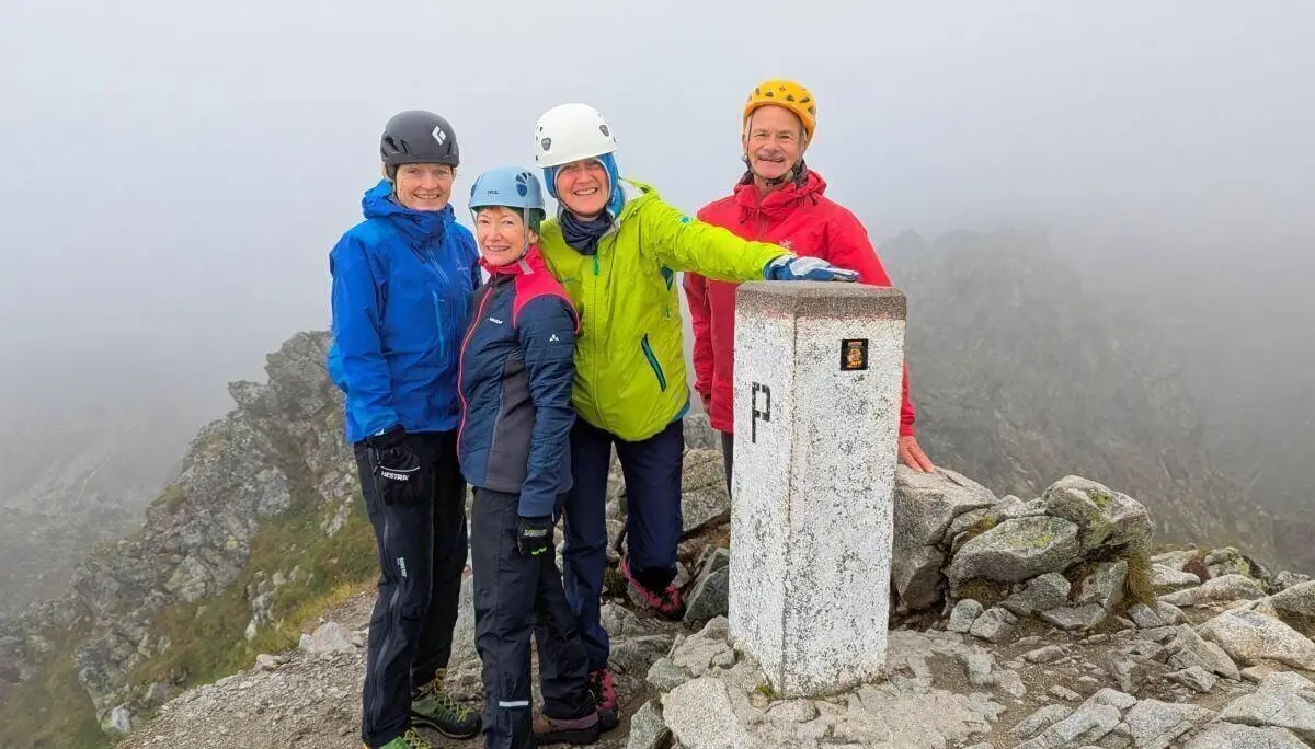 Vier Personen posieren am Grenzstein „P“ auf nebligem Gipfel; sie tragen Helme und Outdoorjacken. | © DAV Markt Schwaben | Foto Michael Huber