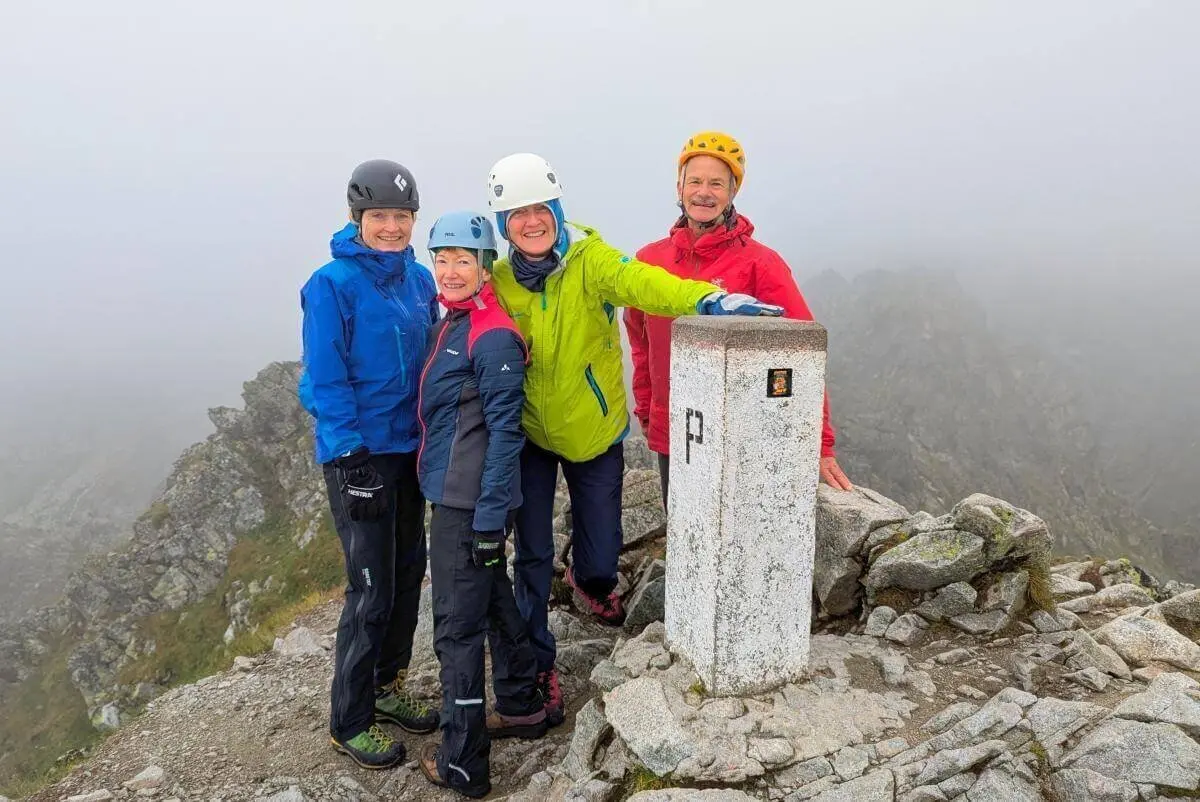 Vier Personen posieren am Grenzstein „P“ auf nebligem Gipfel; sie tragen Helme und Outdoorjacken. | © DAV Markt Schwaben | Foto Michael Huber