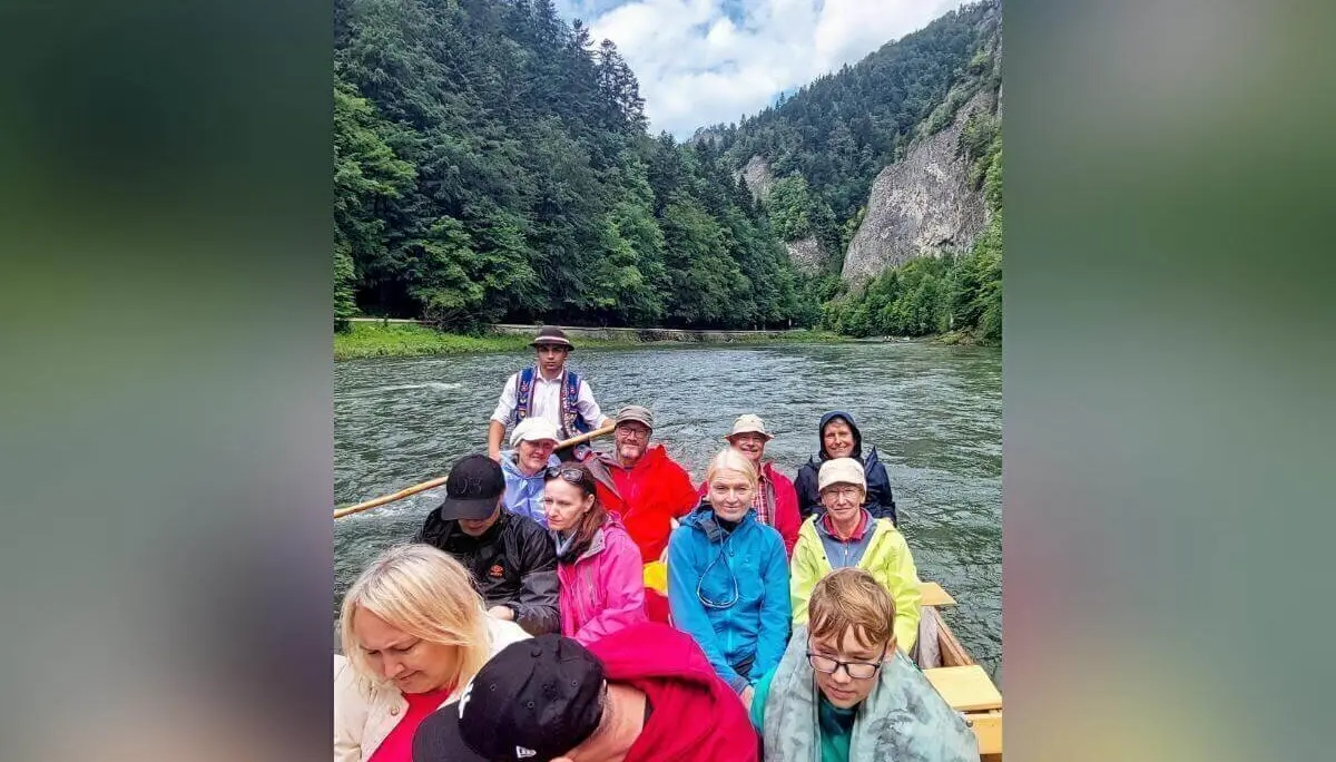 Eine Gruppe fährt in Regenjacken auf Holzboot durch ruhigen Fluss; hinten steht ein Guide in Tracht, umgeben von Wald und Felsen. | © DAV Markt Schwaben | Foto Lutz Gründel