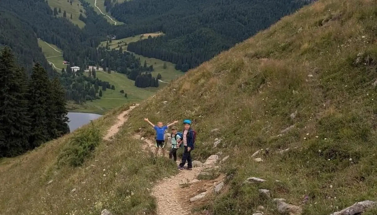 Drei Kinder wandern auf einem Bergpfad bei bewölktem Himmel. Sie gehen durch grünes Gelände mit Sicht auf umliegende Berge. | © DAV Markt Schwaben | Foto Familiengruppe