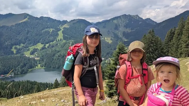 rei Kinder stehen auf einem grasbewachsenen Hang mit Rucksäcken. Im Hintergrund: Wälder, Berge und ein glitzernder See. | © DAV Markt Schwaben | Foto Familiengruppe