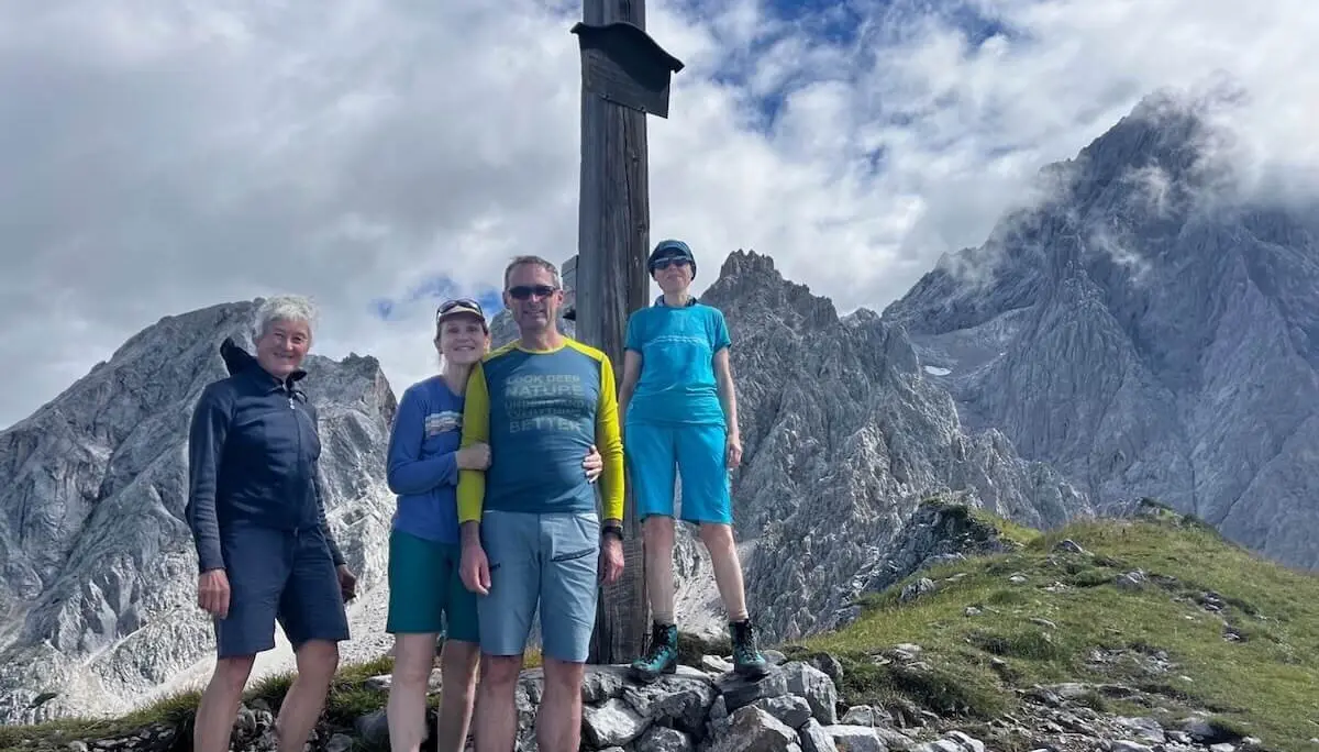 Vier Wandernde posieren vor einem Gipfelkreuz mit Blick auf felsige Bergspitzen unter teils bewölktem Himmel. | © DAV Markt Schwaben | Foto: Markus Sellmeier