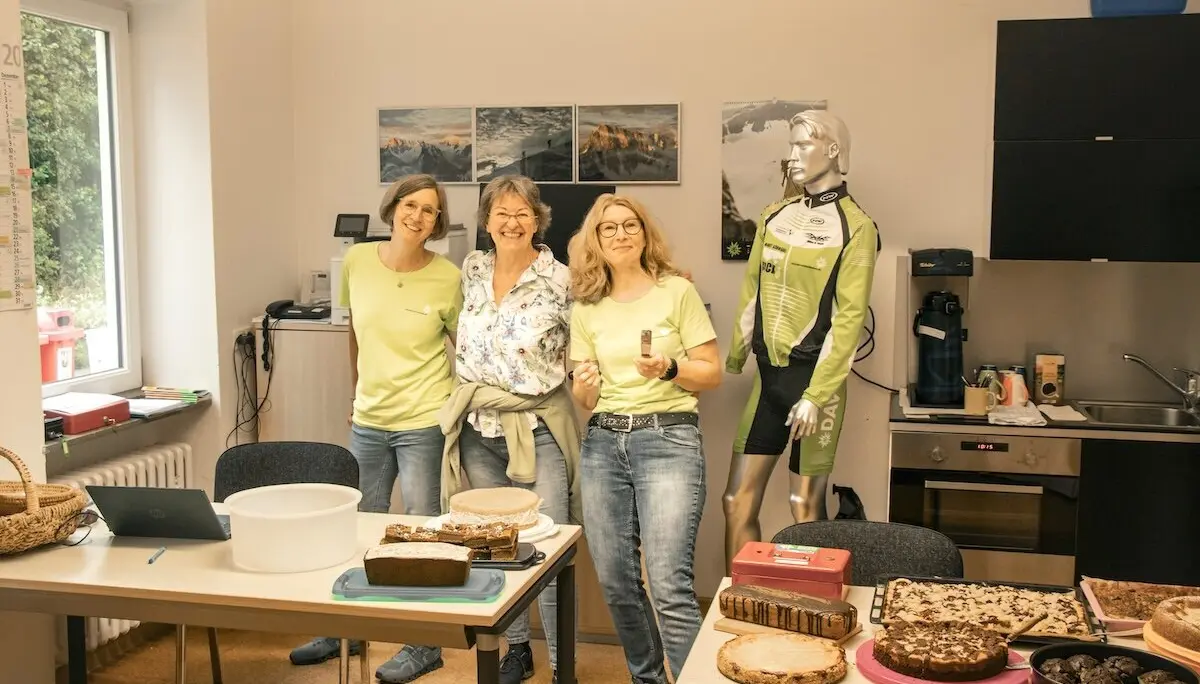 Drei Personen stehen vor einem Tisch mit Kuchen und Gebäck; ein Radtrikot-Mannequin ergänzt die lockere Atmosphäre. | © DAV Markt Schwaben | Foto David Sigl