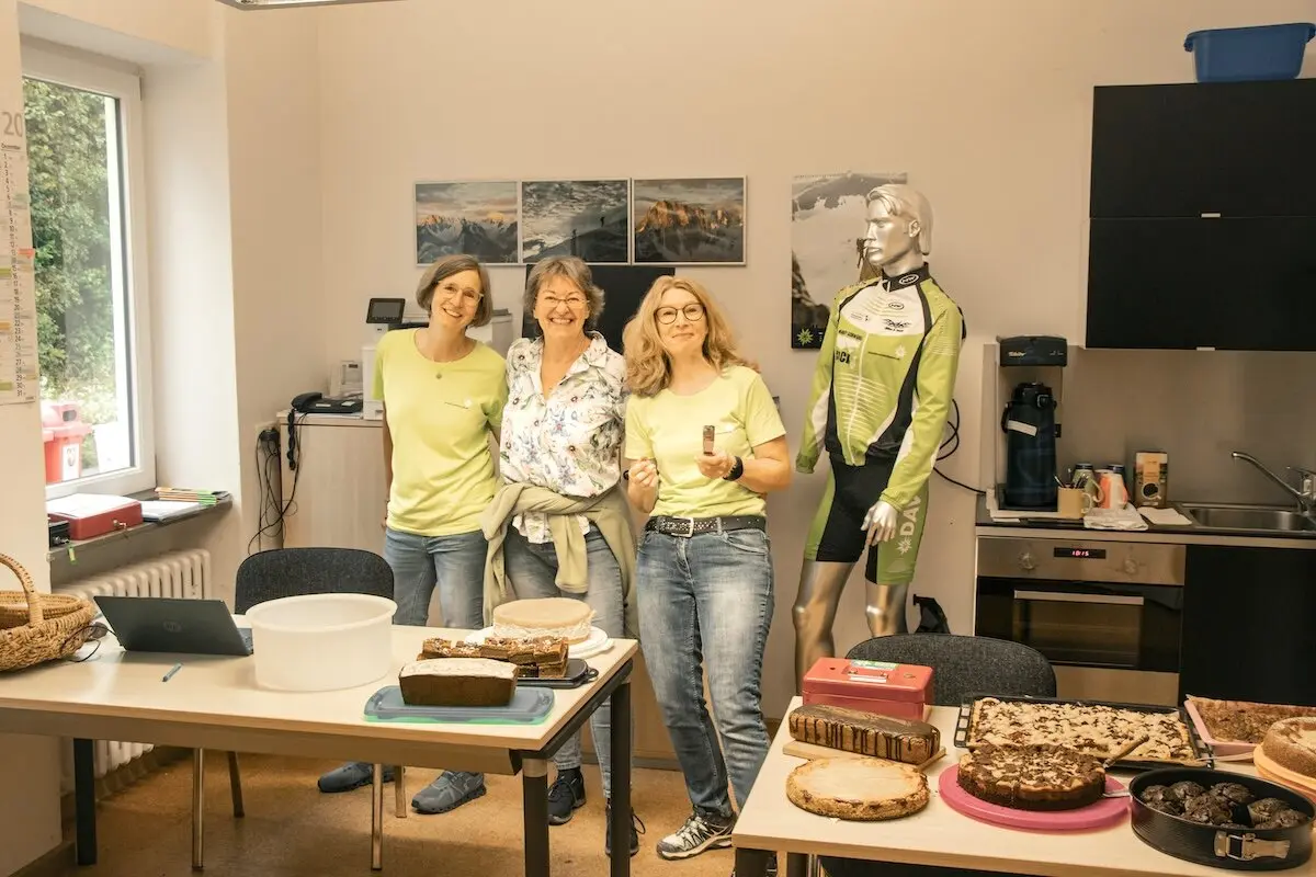 Drei Personen stehen vor einem Tisch mit Kuchen und Gebäck; ein Radtrikot-Mannequin ergänzt die lockere Atmosphäre. | © DAV Markt Schwaben | Foto David Sigl