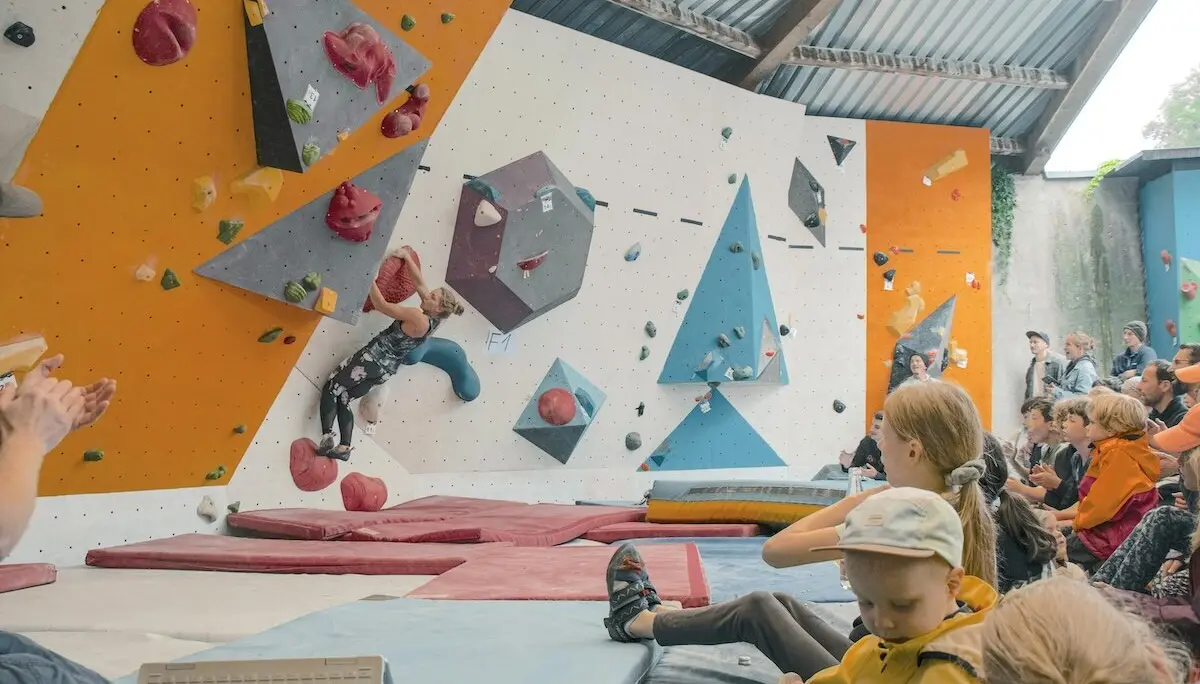Ein Kletterer im Boulderbereich erklimmt die Wand mit roten Griffen, Zuschauer sitzen und stehen gespannt daneben. | © DAV Markt Schwaben | Foto David Sigl