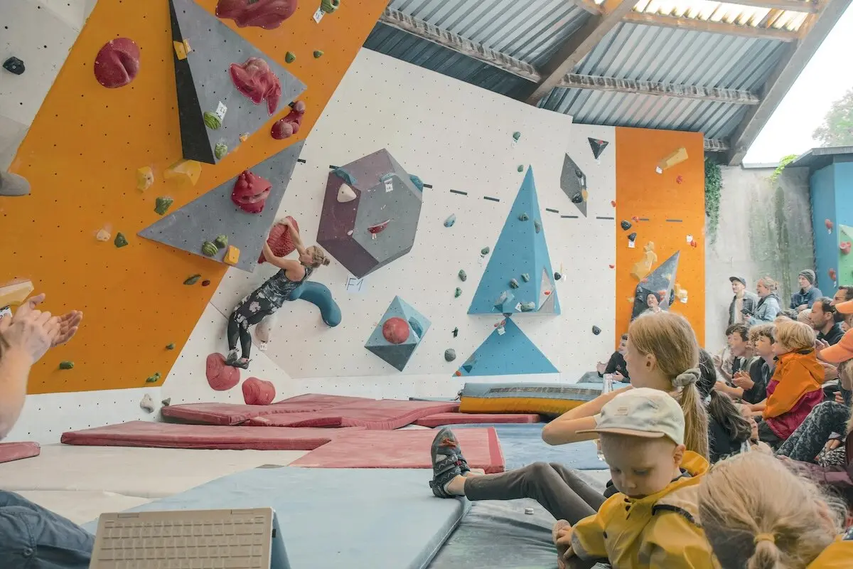 Ein Kletterer im Boulderbereich erklimmt die Wand mit roten Griffen, Zuschauer sitzen und stehen gespannt daneben. | © DAV Markt Schwaben | Foto David Sigl