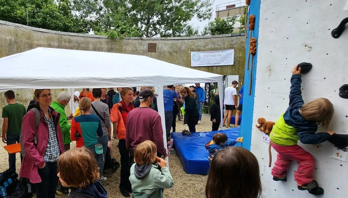 Zwei Kinder klettern gesichert an der Outdoor-Wand, umgeben von Besucher*innen unter einem weißen Pavillon. | © DAV Markt Schwaben | Foto Anna Thoms