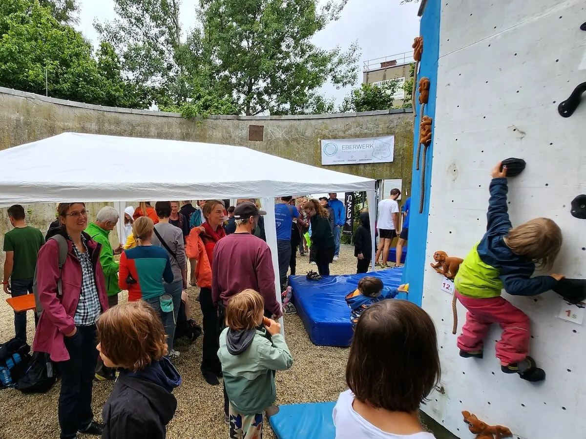 Zwei Kinder klettern gesichert an der Outdoor-Wand, umgeben von Besucher*innen unter einem weißen Pavillon. | © DAV Markt Schwaben | Foto Anna Thoms