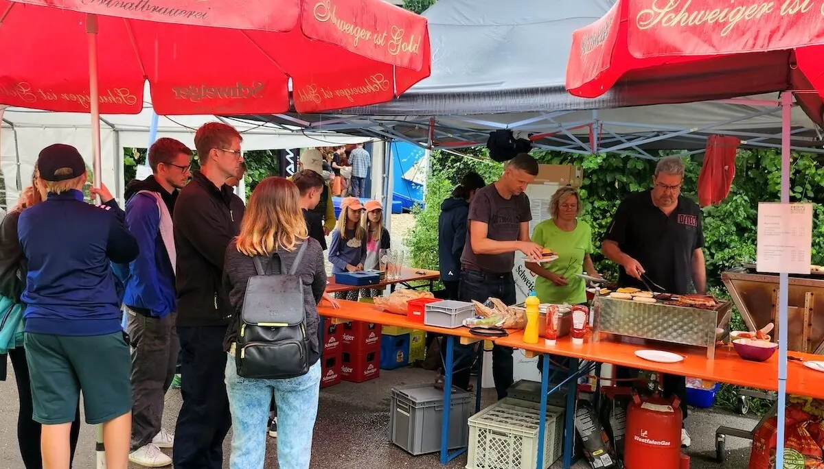 Besucher*innen stehen am Grillstand mit roten Schweiger-Schirmen, während Helfer Essen ausgeben – gesellige Stimmung im Freien. | © DAV Markt Schwaben | Foto Anna Thoms