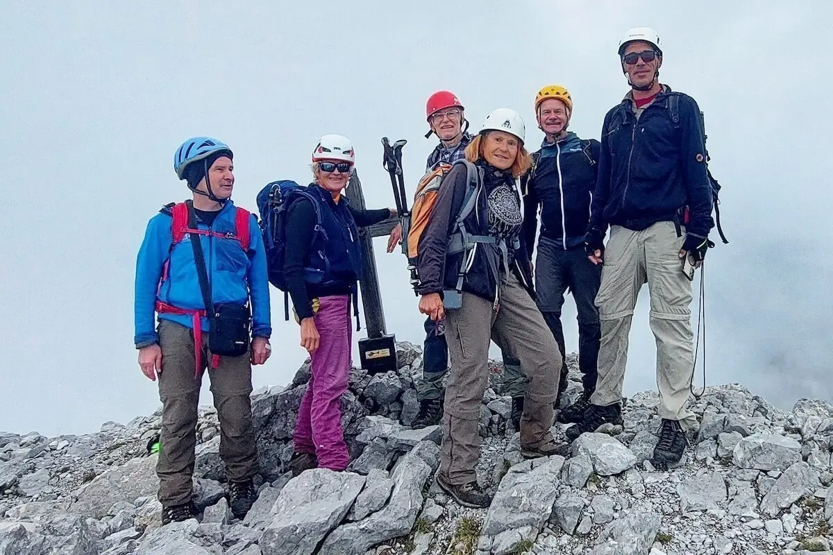 Sechs Bergsteiger stehen am Gipfelkreuz auf felsigem Grat, im Hintergrund Wolken und Bergstimmung. | © DAV Markt Schwaben · Foto: Lutz Gründel