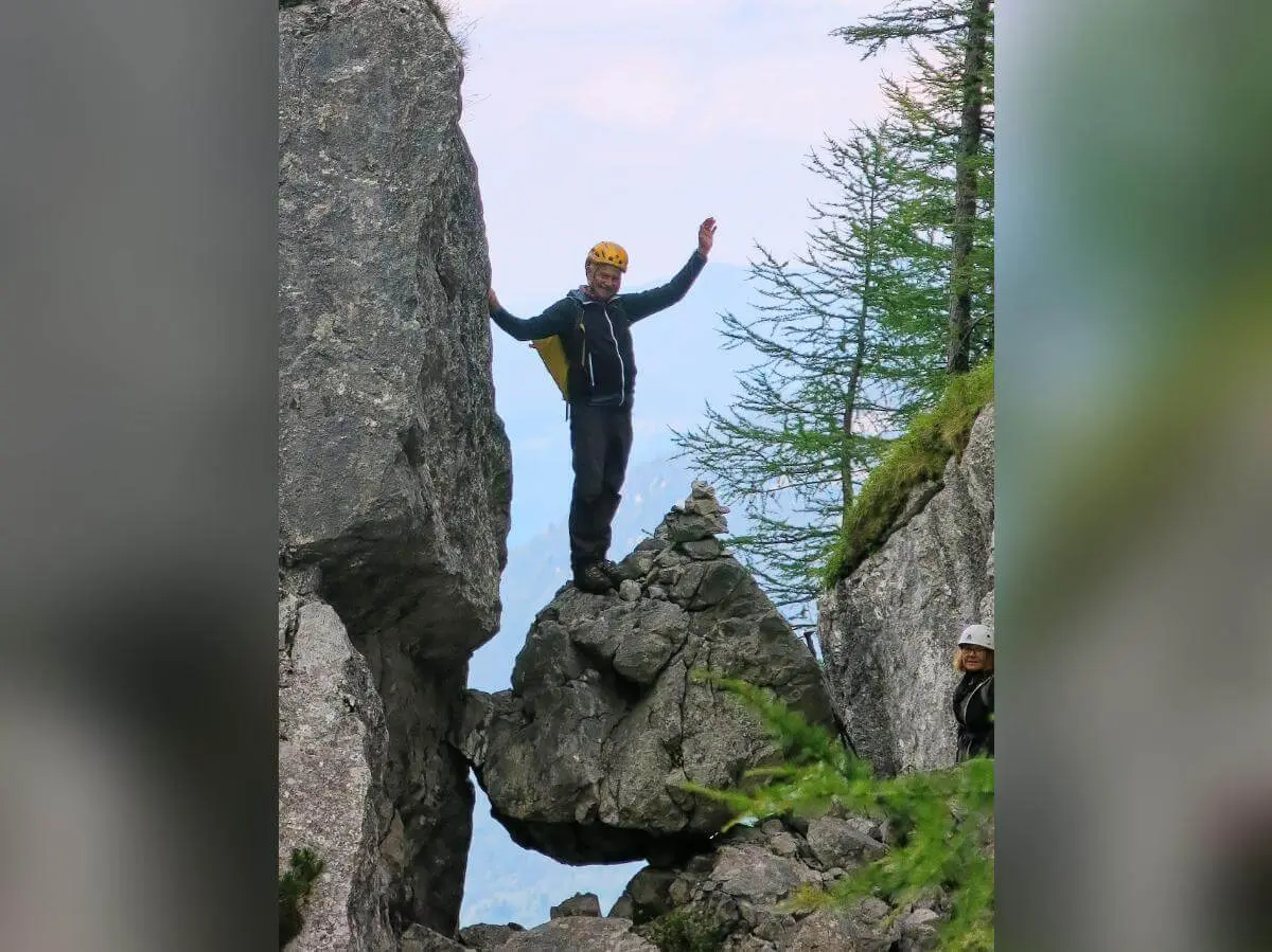 Ein Bergsteiger steht mit erhobenen Armen auf einem Felsblock im Fensterl, hinten Wald und Bergpanorama. | © DAV Markt Schwaben · Foto: Erwin Matzinger