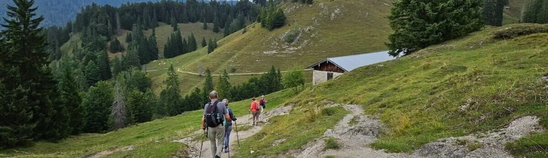 Drei Wandernde mit Rucksäcken und Stöcken auf einem Bergpfad, im Hintergrund eine Holzhütte und bewaldete Hänge. | © DAV Markt Schwaben | Foto Hubert Inhofer