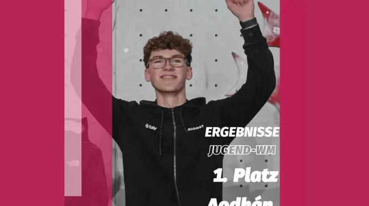 Aodhán Umlauf steht stolz vor einer Kletterwand, die Arme siegreich erhoben. Er trägt eine schwarze Sportjacke mit den Logos von DAV und Mammut. Im Bild ist der Text ‚1. Platz Jugend-WM‘ zu sehen. Das Foto feiert seinen Sieg bei der Jugend-Weltmeisterschaft im Sportklettern. | © iFSC Instagram: @ifsclimbing