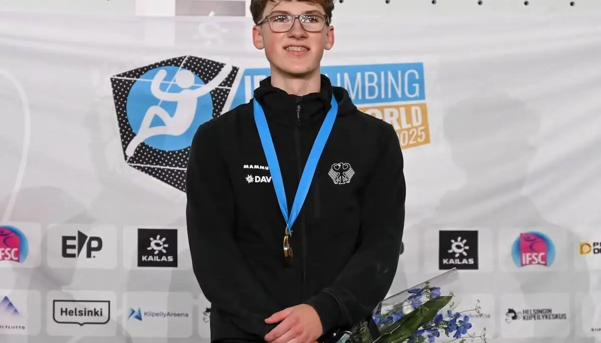 Aodhán Umlauf, 18, steht bei der U19‑WM‑Siegerehrung in Helsinki mit Goldmedaille und Blumenstrauß vor einer Sponsorwand. | © Foto: Richard Aspland / IFSC