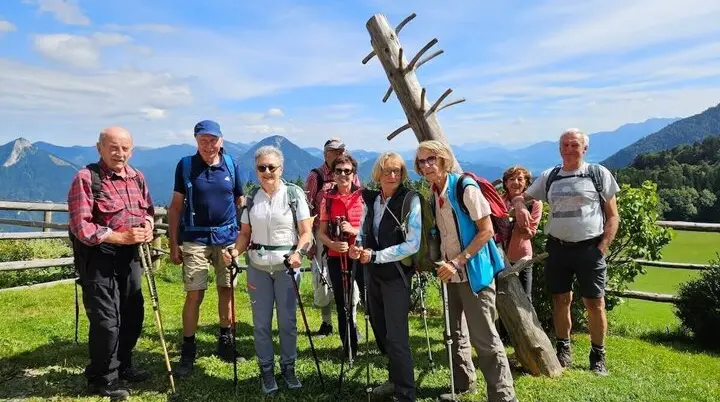 Neun Wandernde stehen auf Wiese vor Holzskulptur und Zaun, dahinter Hügel und Berglandschaft bei Sonnenschein. | © DAV Markt Schwaben | Foto Hubert Inhofer