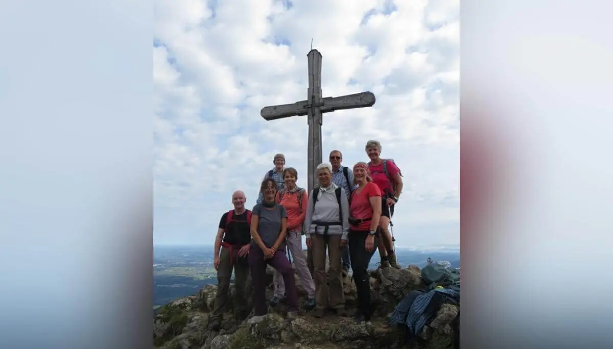Neun Wandernde stehen am Gipfelkreuz auf felsigem Bergplateau; im Hintergrund wolkiger Himmel und weite Aussicht. | © DAV Markt Schwaben | Foto Erwin Matzinger