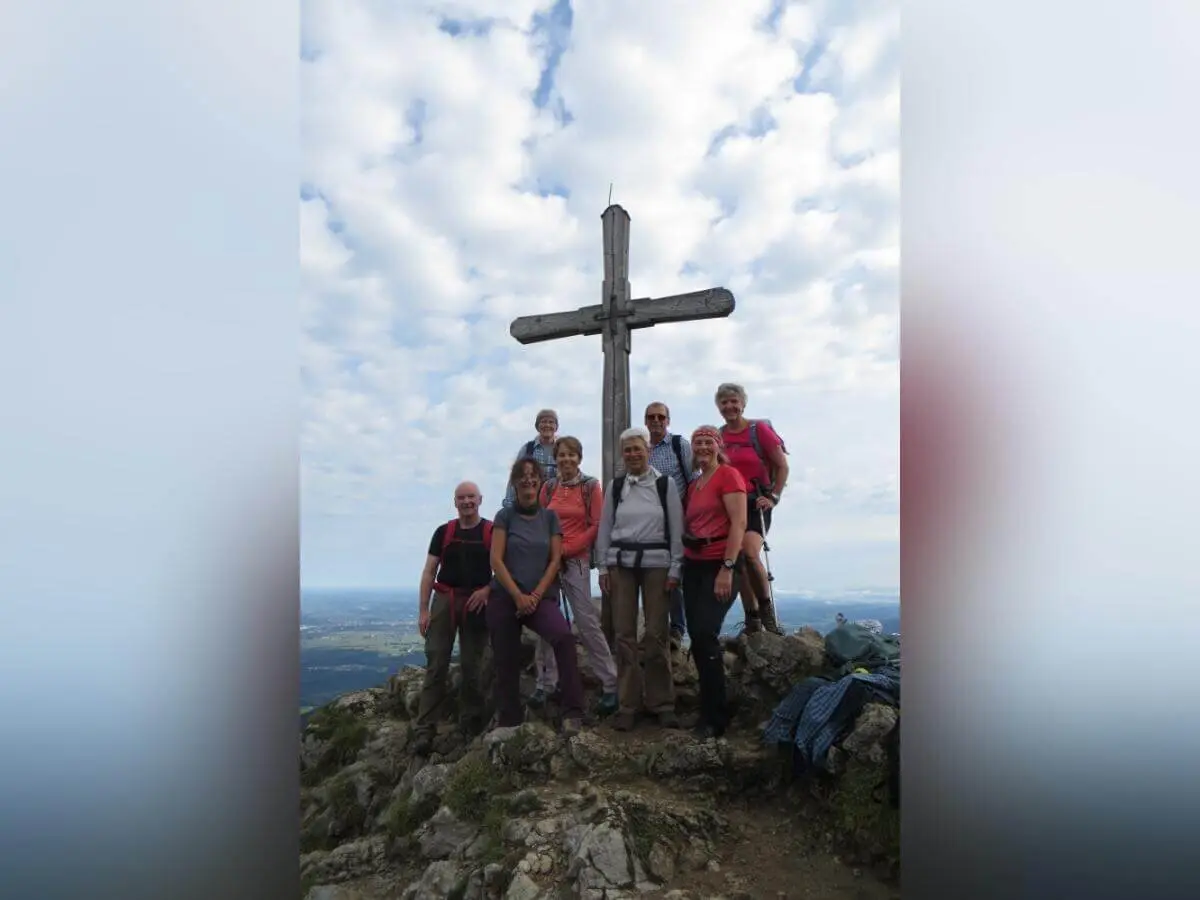 Neun Wandernde stehen am Gipfelkreuz auf felsigem Bergplateau; im Hintergrund wolkiger Himmel und weite Aussicht. | © DAV Markt Schwaben | Foto Erwin Matzinger