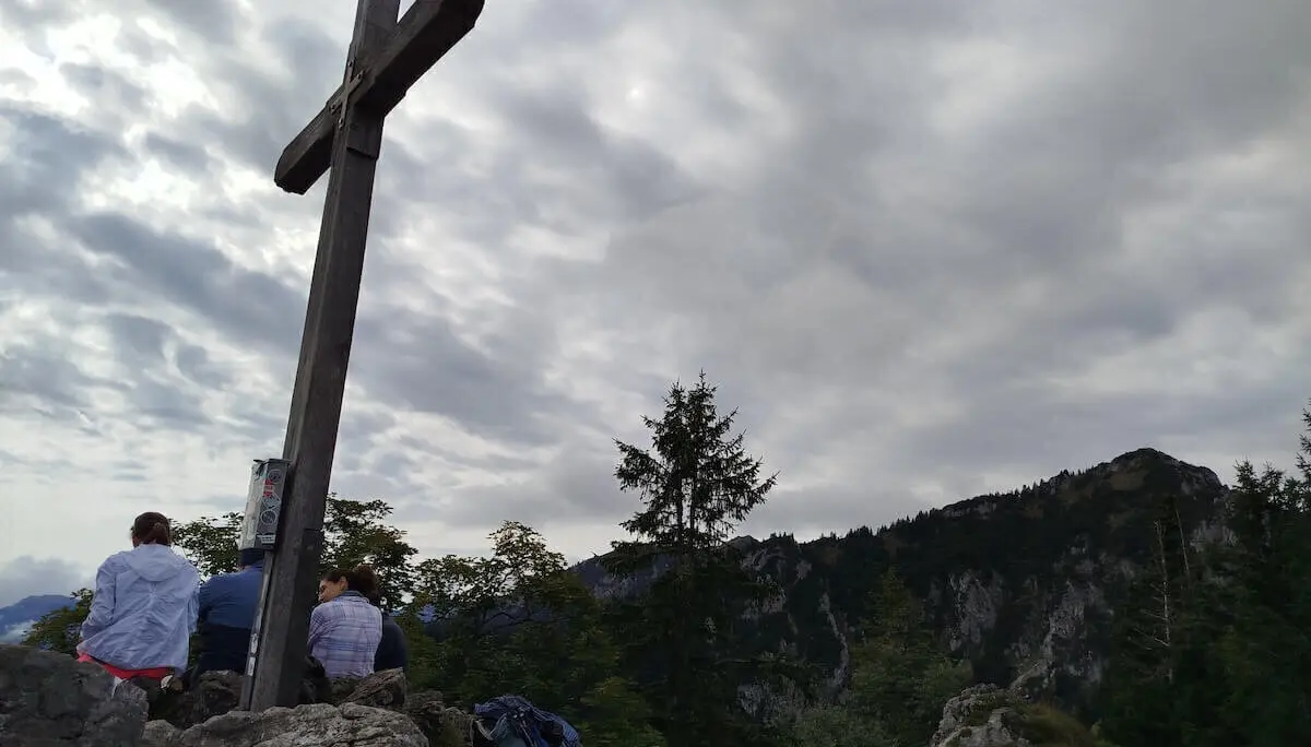 Mehrere Wandernde sitzen am hölzernen Gipfelkreuz auf Felsen; im Hintergrund bewaldete Berge unter wolkigem Himmel. | © DAV Markt Schwaben | Foto Erwin Matzinger