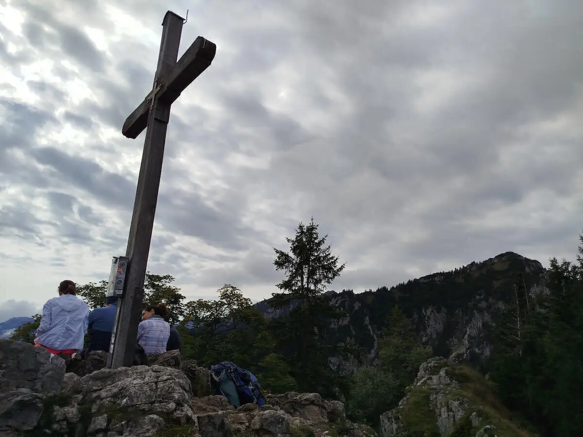 Mehrere Wandernde sitzen am hölzernen Gipfelkreuz auf Felsen; im Hintergrund bewaldete Berge unter wolkigem Himmel. | © DAV Markt Schwaben | Foto Erwin Matzinger