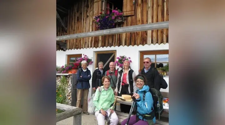 Acht Wandernde stehen vor der Rinner Alm mit Holzfassade und Blumen; eine Person sitzt mit Käseteller auf der Bank. | © DAV Markt Schwaben | Foto Erwin Matzinger