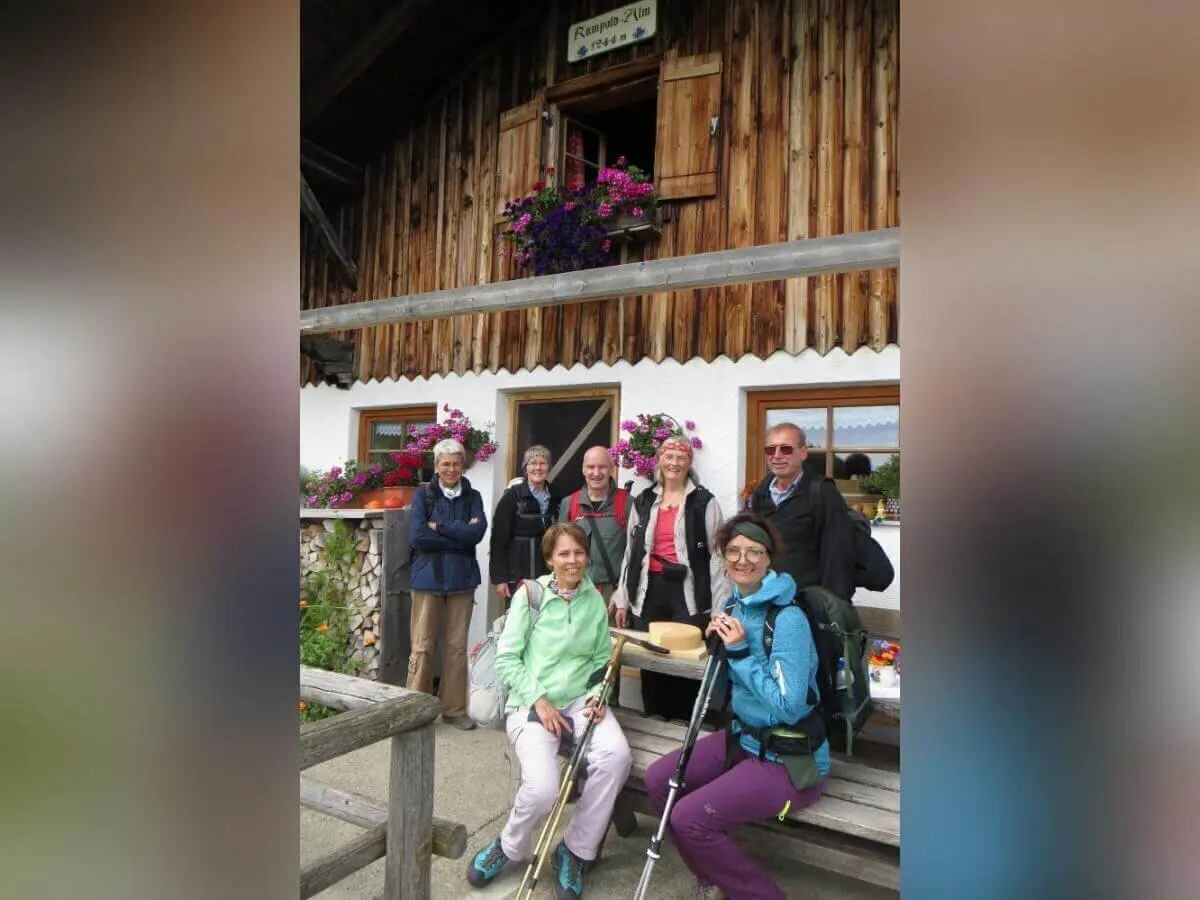 Acht Wandernde stehen vor der Rinner Alm mit Holzfassade und Blumen; eine Person sitzt mit Käseteller auf der Bank. | © DAV Markt Schwaben | Foto Erwin Matzinger