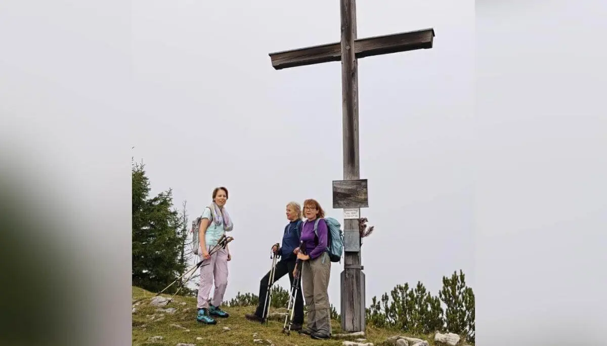 Drei Wandernde stehen mit Stöcken neben einem großen Holzkreuz auf felsigem Grat; bewölkter Himmel im Hintergrund. | © DAV Markt Schwaben | Foto Helga Peters