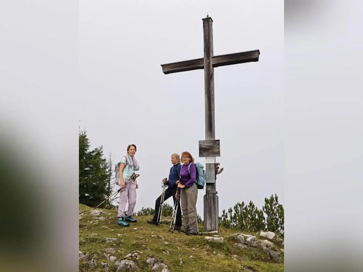 Drei Wandernde stehen mit Stöcken neben einem großen Holzkreuz auf felsigem Grat; bewölkter Himmel im Hintergrund. | © DAV Markt Schwaben | Foto Helga Peters