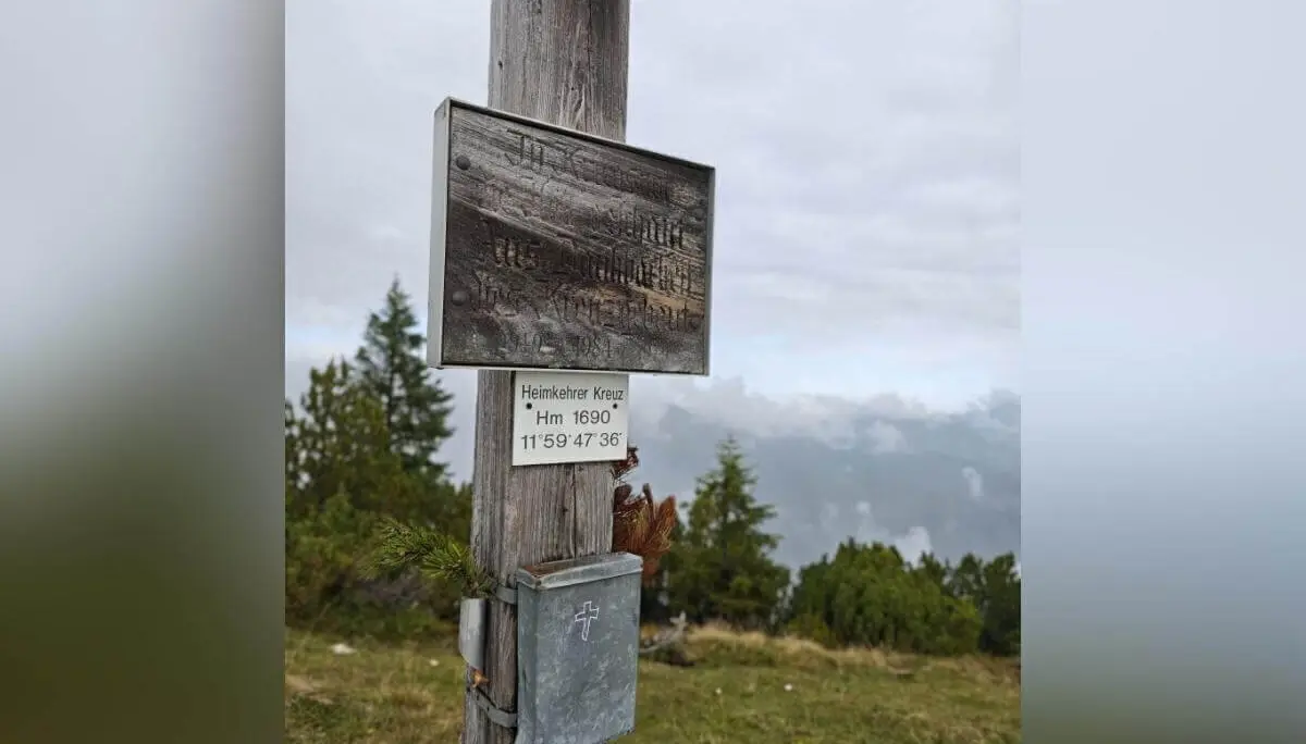 Holzpfosten mit zwei Metallplaketten auf Almwiese; Hintergrund mit Bäumen und wolkenverhangenem Himmel. | © DAV Markt Schwaben | Foto Helga Peters