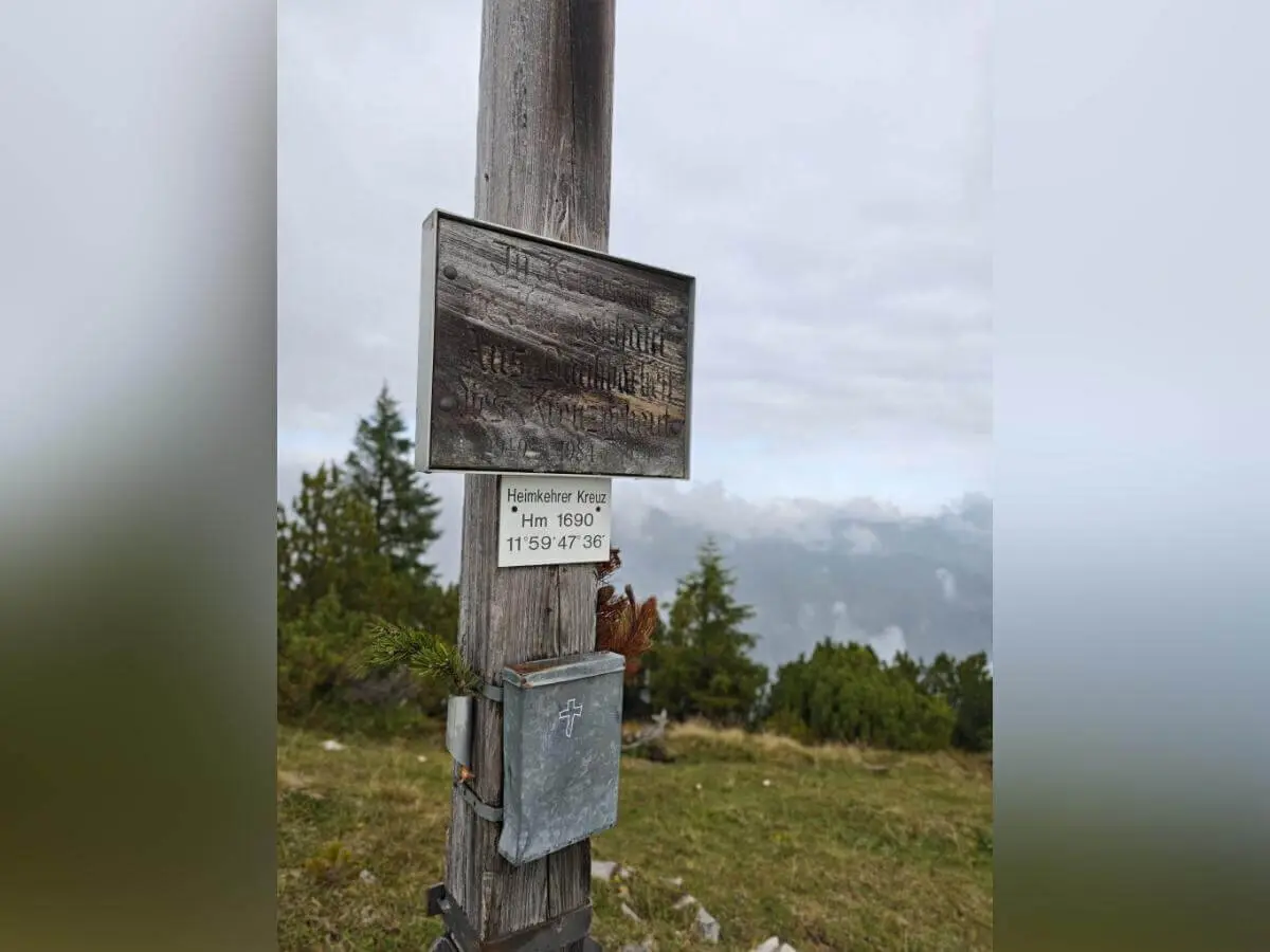 Holzpfosten mit zwei Metallplaketten auf Almwiese; Hintergrund mit Bäumen und wolkenverhangenem Himmel. | © DAV Markt Schwaben | Foto Helga Peters