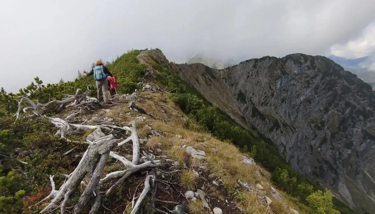 Zwei Wandernde auf schmalem Grat vor steiler Felswand; Wolken hängen tief über dem felsigen Berghang. | © DAV Markt Schwaben | Foto Helga Peters