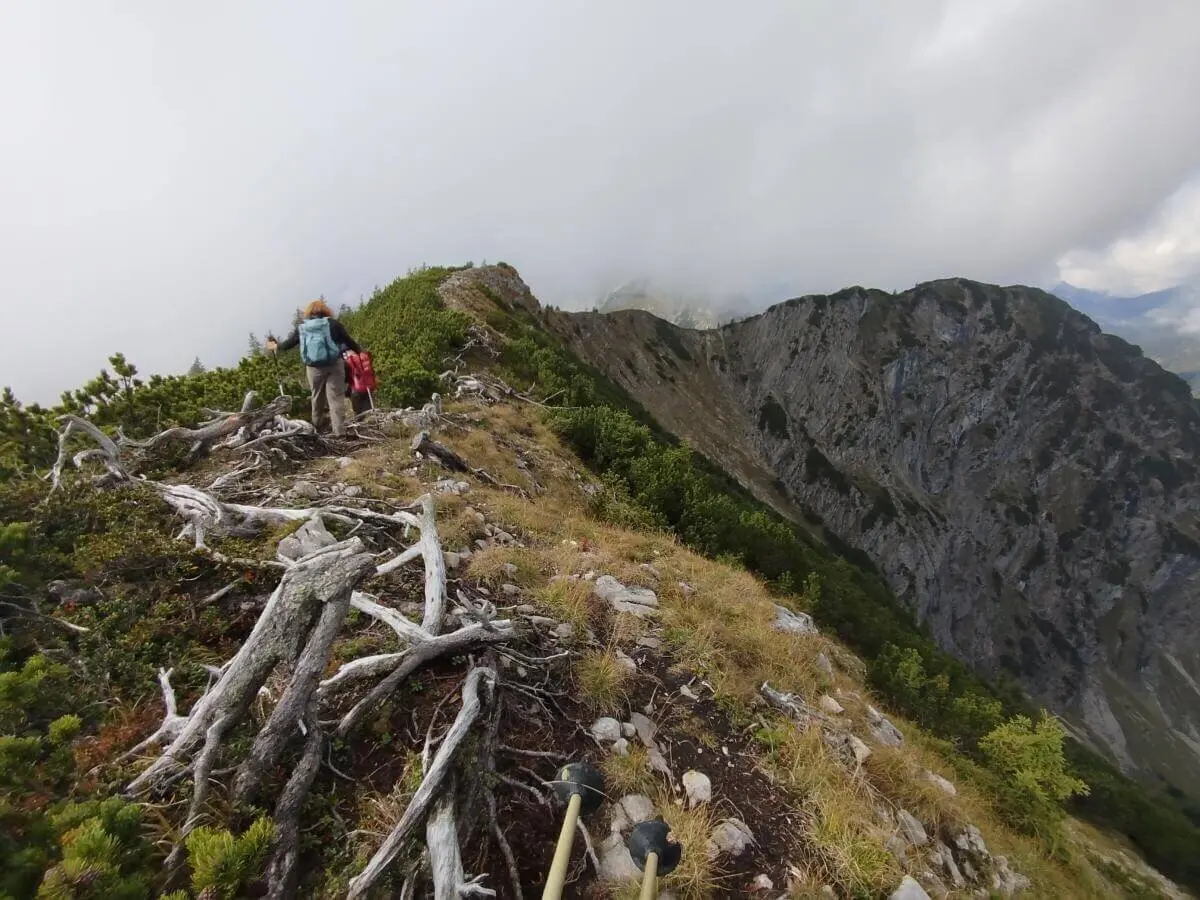 Zwei Wandernde auf schmalem Grat vor steiler Felswand; Wolken hängen tief über dem felsigen Berghang. | © DAV Markt Schwaben | Foto Helga Peters