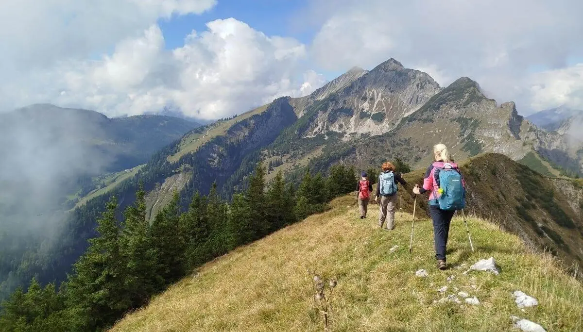 Drei Wandernde stehen vor rustikaler Almhütte aus Holz und Stein; hügelige Landschaft und Wolken im Hintergrund. | © DAV Markt Schwaben | Foto Helga Peters