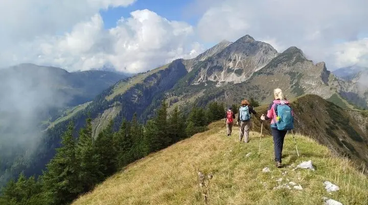 Drei Wandernde stehen vor rustikaler Almhütte aus Holz und Stein; hügelige Landschaft und Wolken im Hintergrund. | © DAV Markt Schwaben | Foto Helga Peters